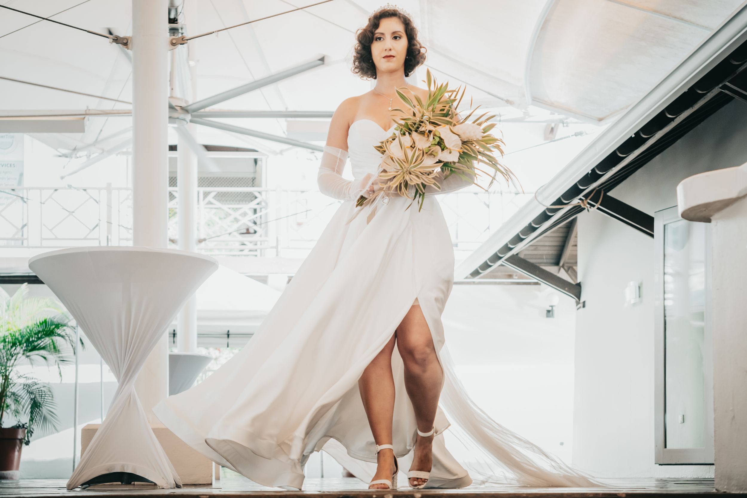 Bride in flowing white dress holding dried floral bouquet poses in elegant indoor venue setting.