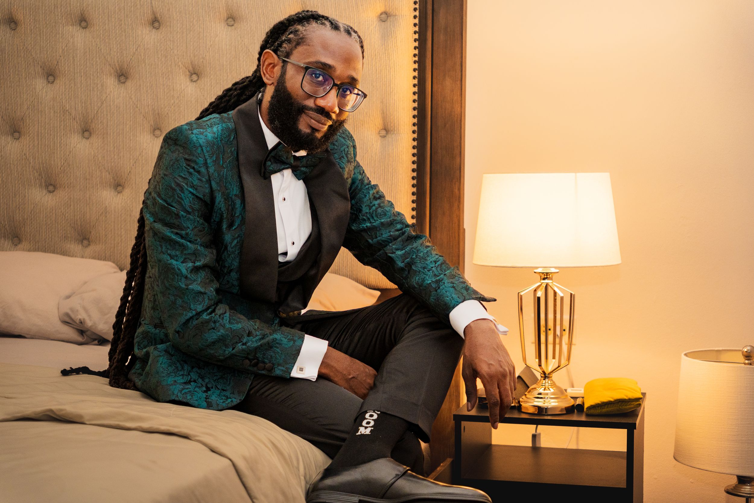 Formal wear in emerald green velvet tuxedo jacket seated on beige tufted furniture.