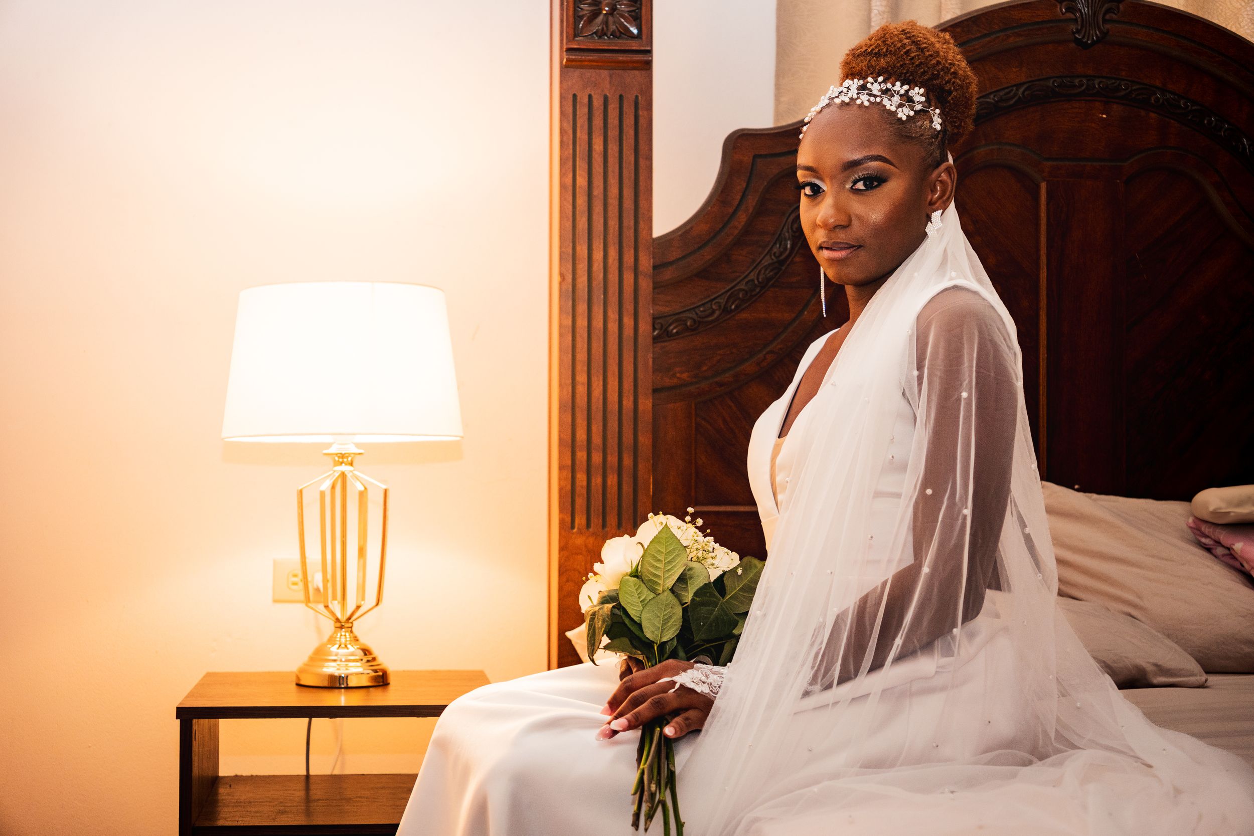 Bridal portrait in classic white gown and veil against warm-lit bedroom setting.