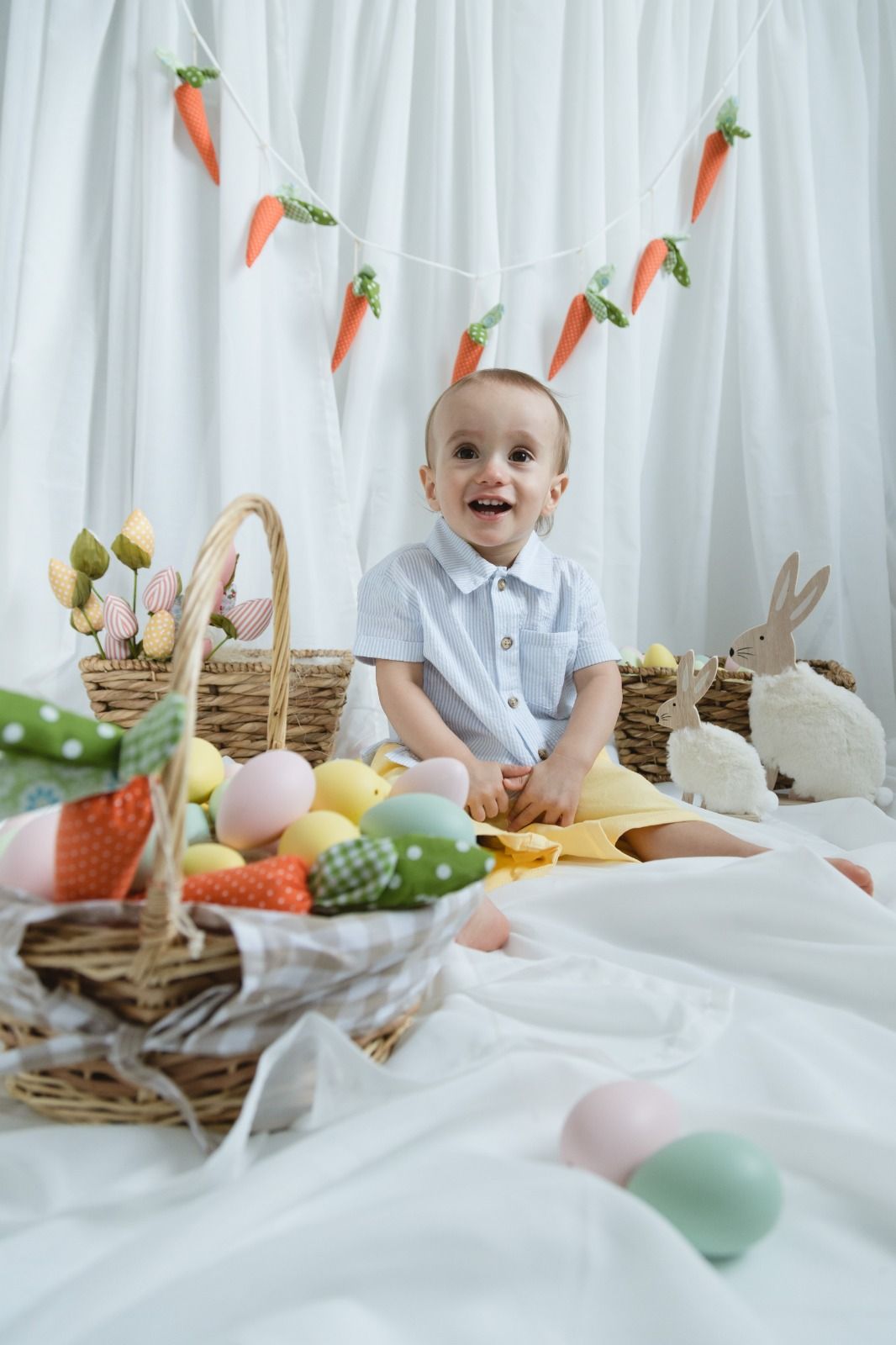Easter holiday scene with baskets, decorated eggs, carrot garland, and a young child in white sitting amid festive decor.