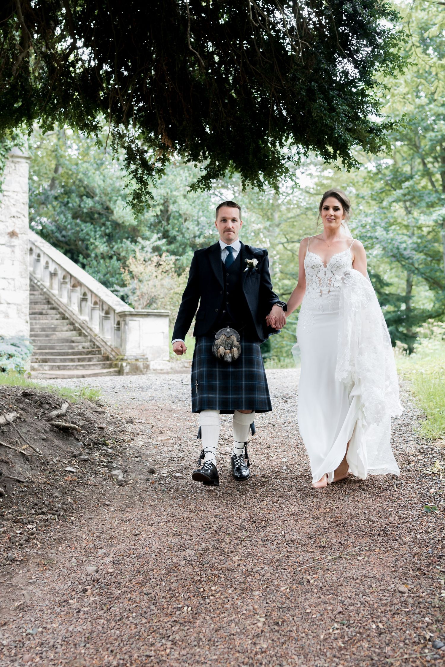 Newlyweds walk together on a garden path with groom wearing traditional Scottish kilt and bride in elegant white gown.