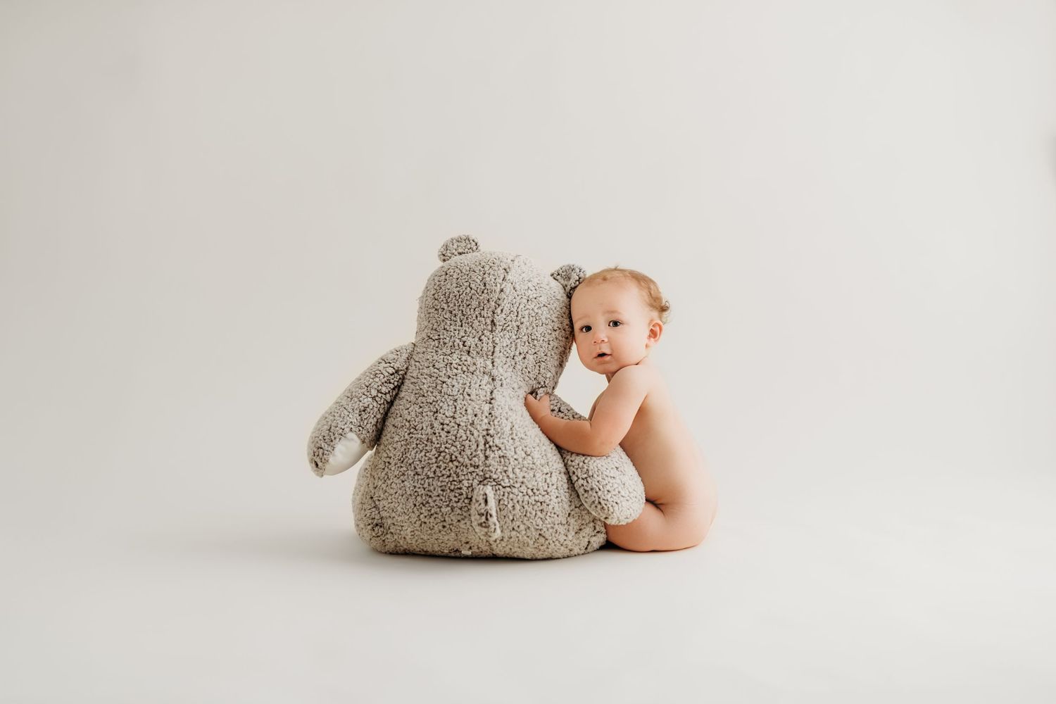 Baby cuddling with large grey teddy bear against white background.