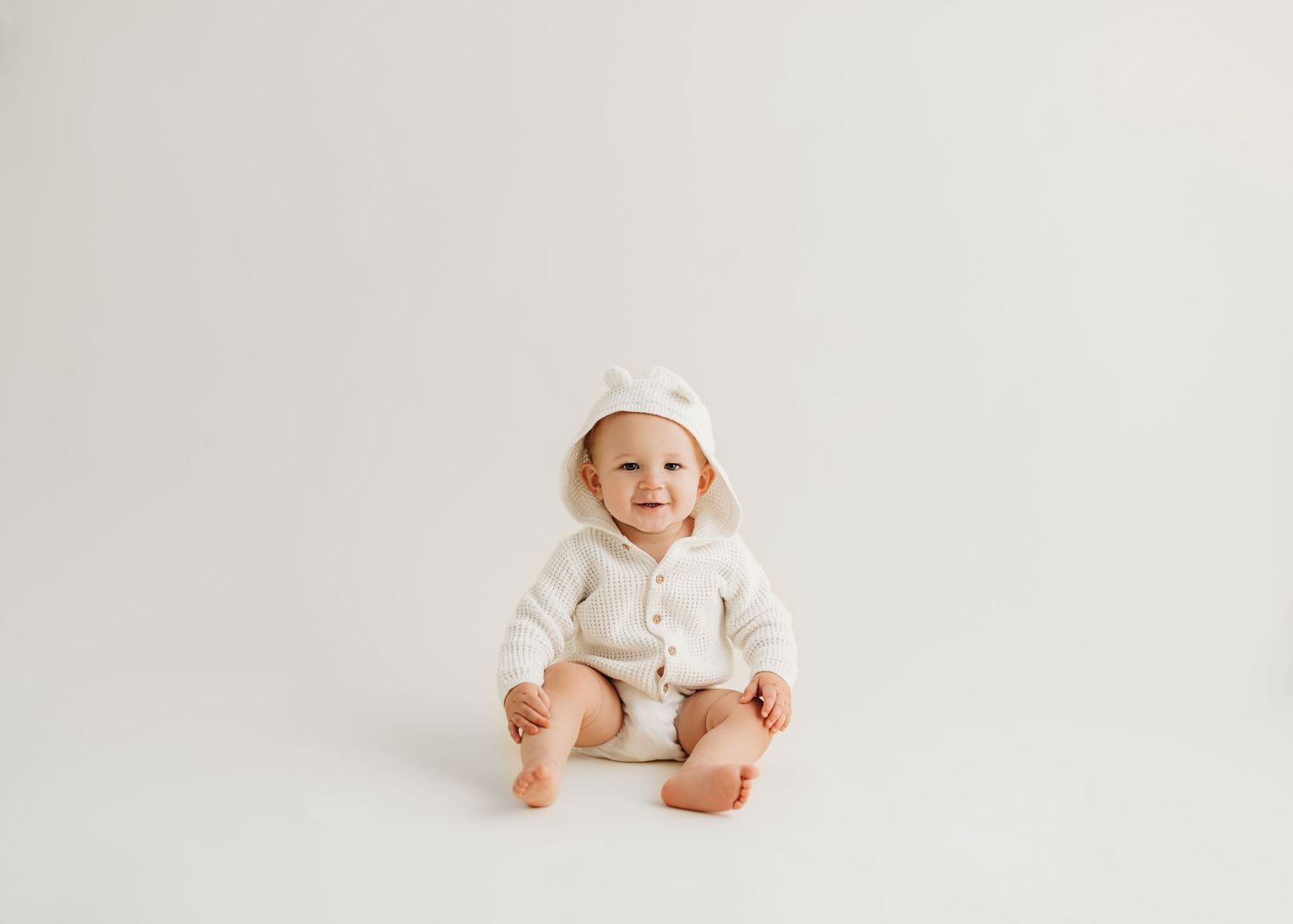 Baby wearing white sweater sits on neutral background, giggling with joy.