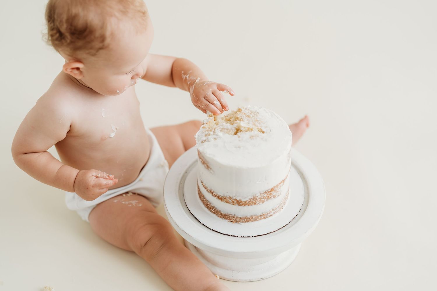 Baby in diaper reaching for white tiered cake against plain backdrop.