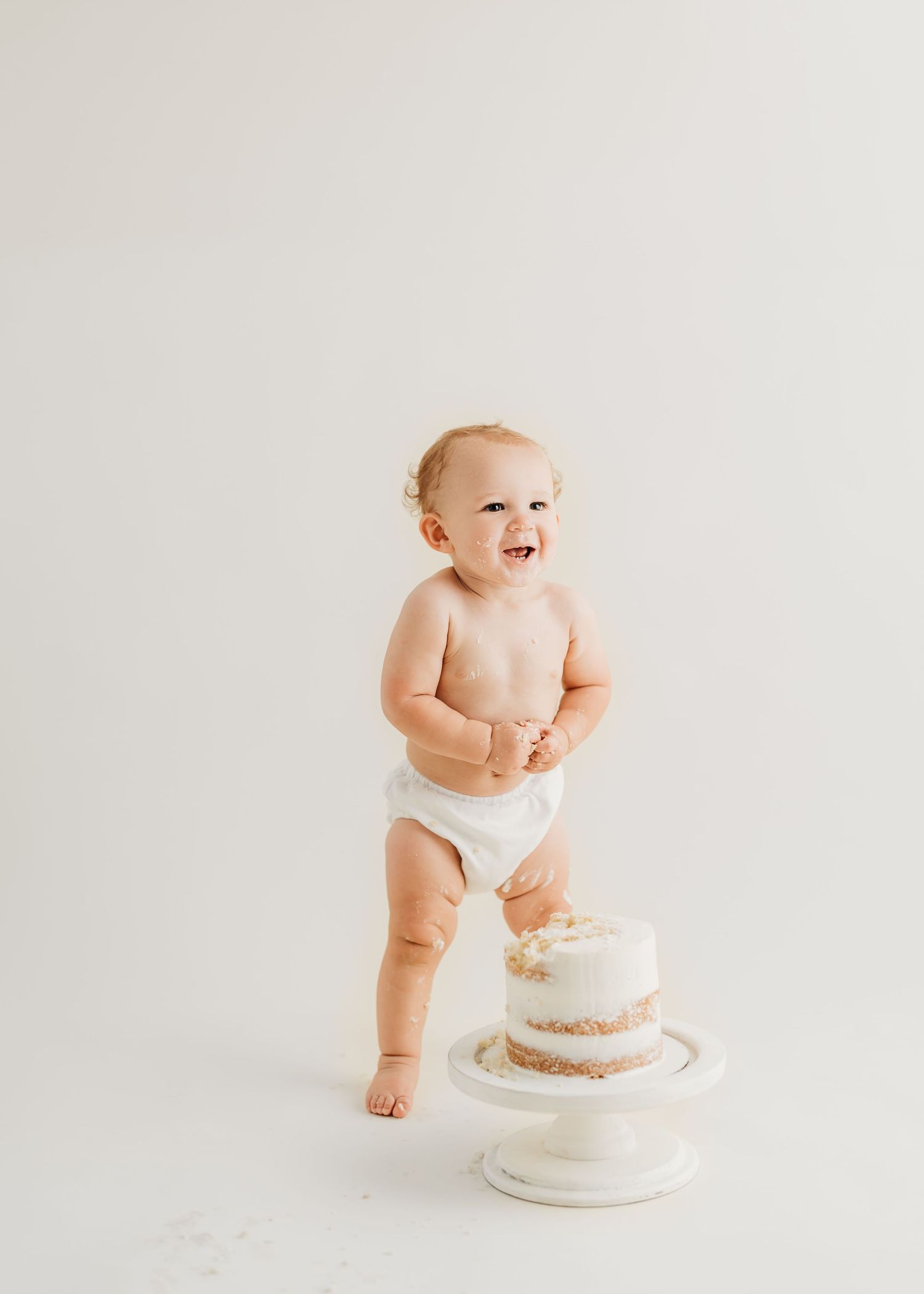 Joyful baby in diaper standing next to white decorated cake on minimalist background.