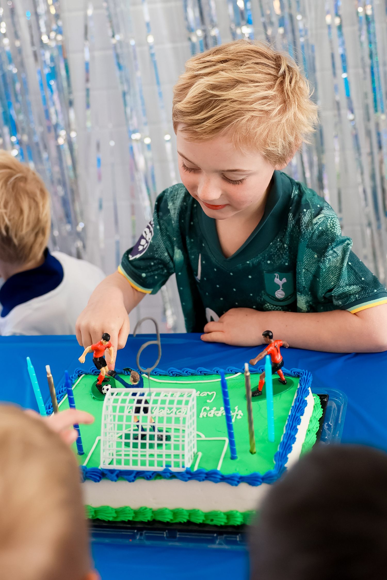 Child in green plays with a birthday cake decorated like a soccer field with blue frosting and plastic figures on top.