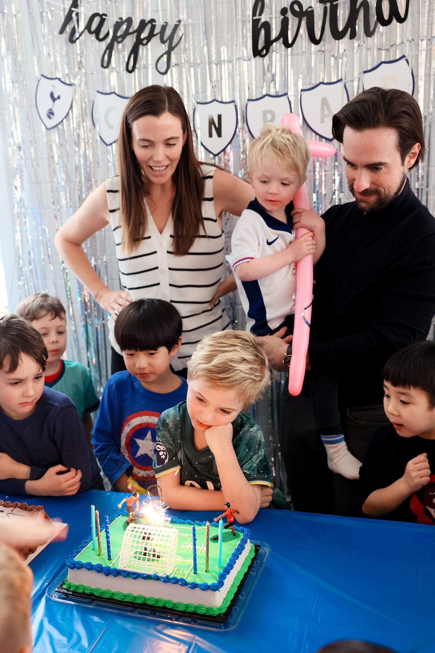 A group gathers around a bright blue and green birthday cake at a party with silver decorations in the background.