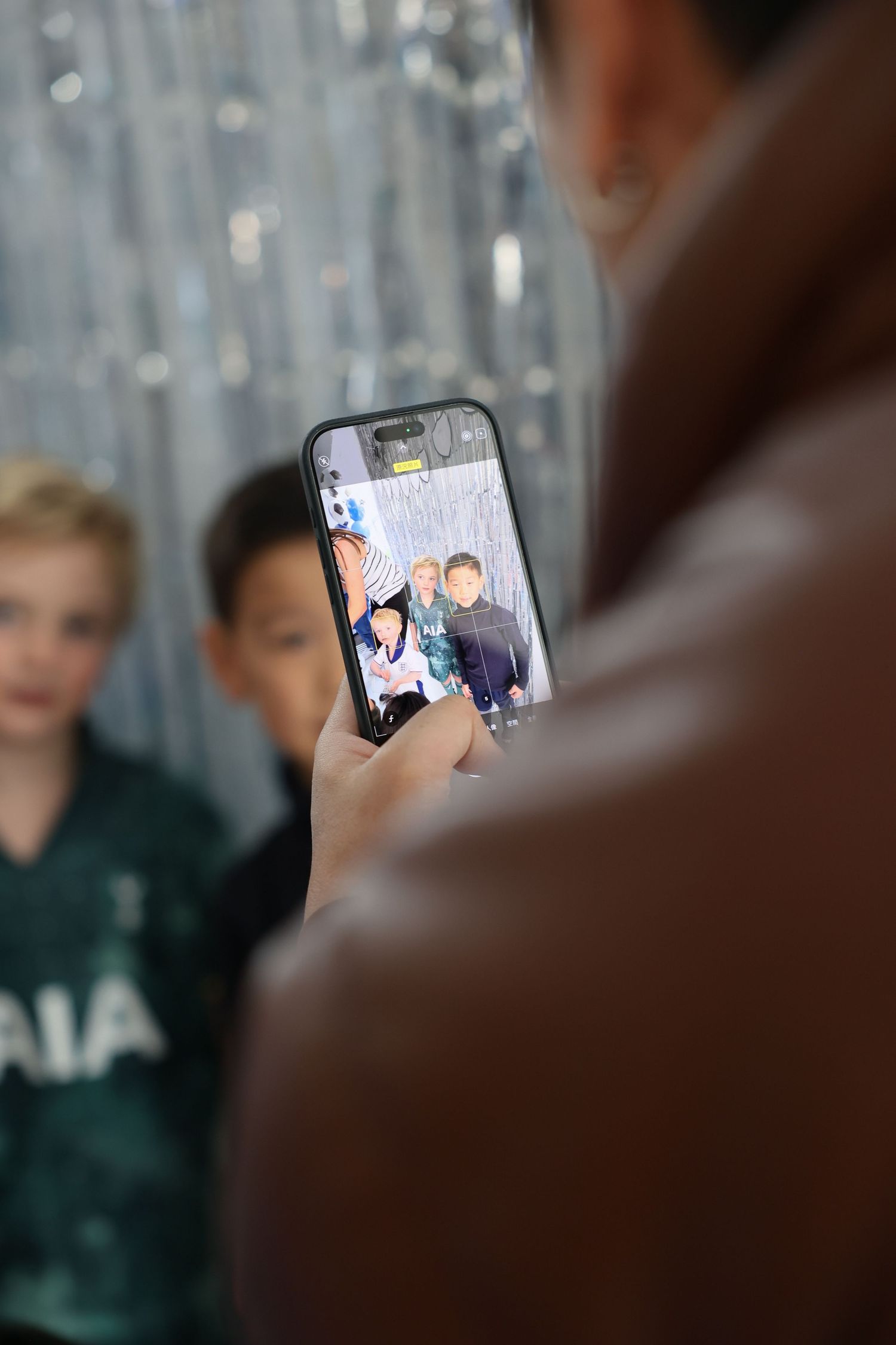 Someone holds a smartphone showing a photo on screen while wearing a Tottenham Hotspur jersey in the background. Smilein Photos