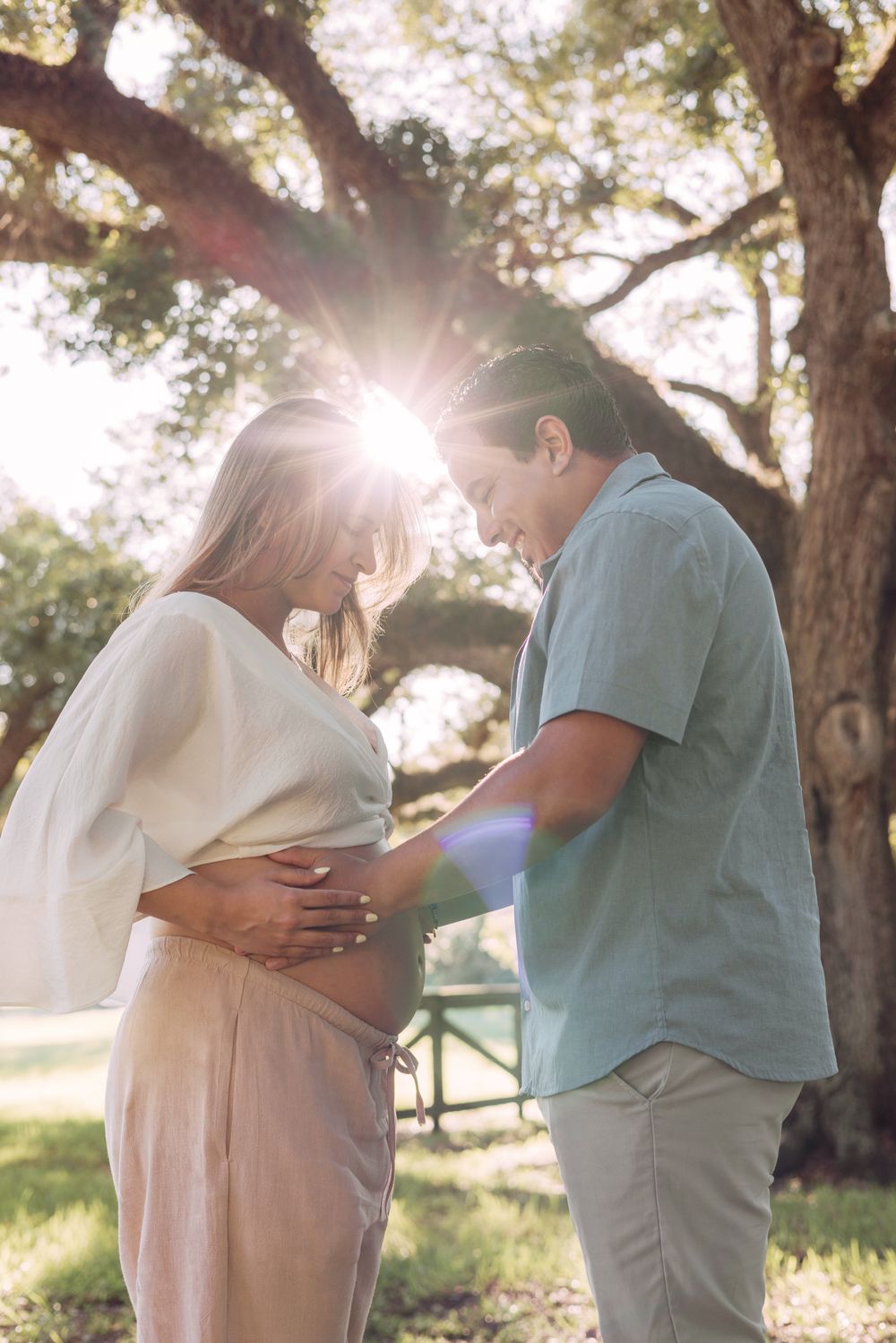 Intimate pregnancy photos of couple embracing in sunlit park with trees in background.