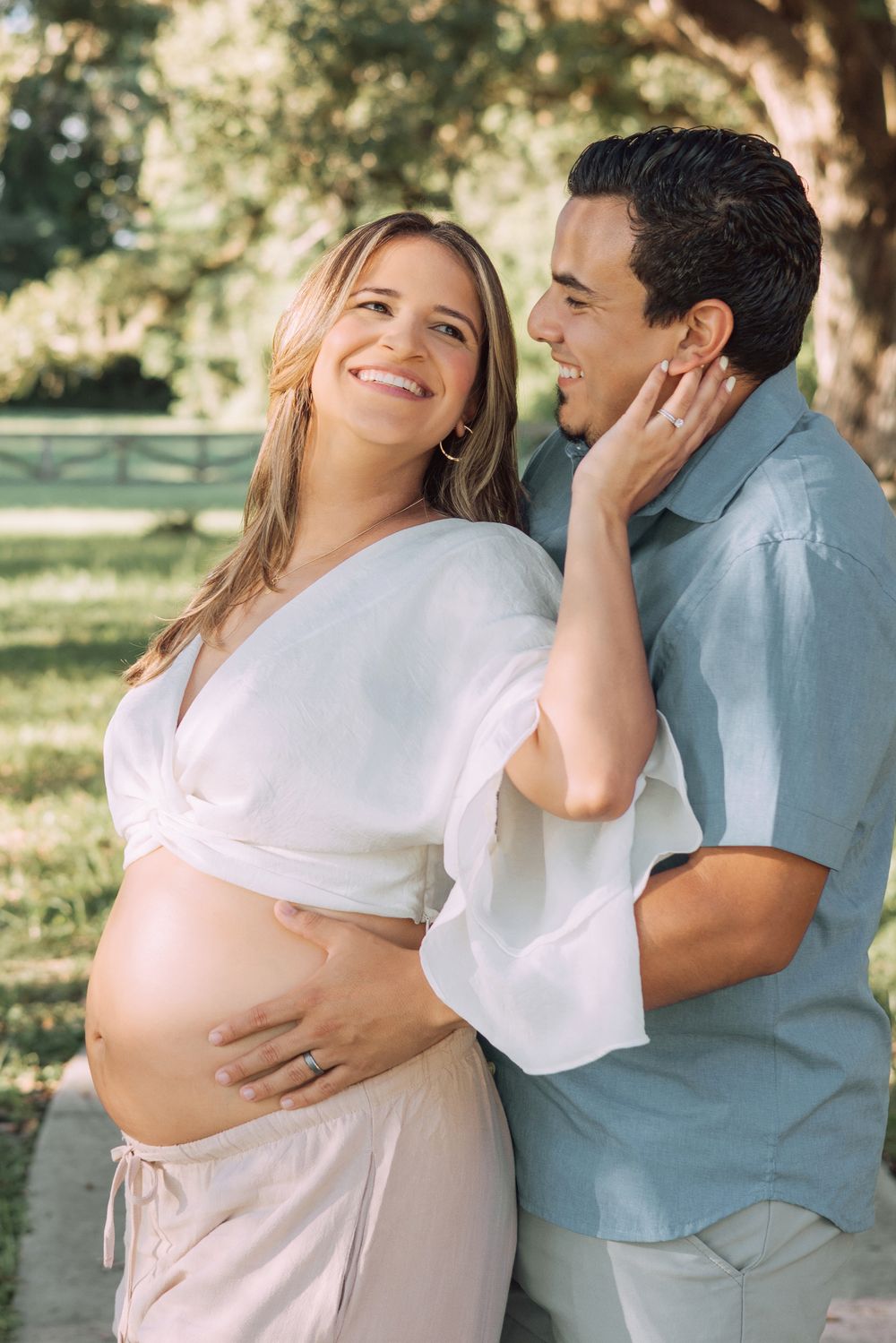 Romantic maternity photoshoot showing tender moments between expecting couple in outdoor setting with sunlit trees.