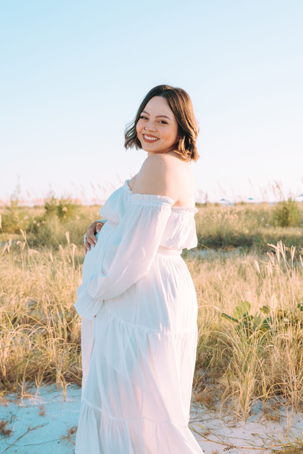 A dreamy maternity photoshoot on a sandy beach at sunset with soft white flowing dress and natural lighting.