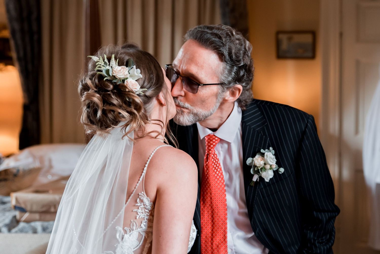 A bride in a lace wedding dress with veil shares a tender moment with a suited guest wearing glasses and red tie.