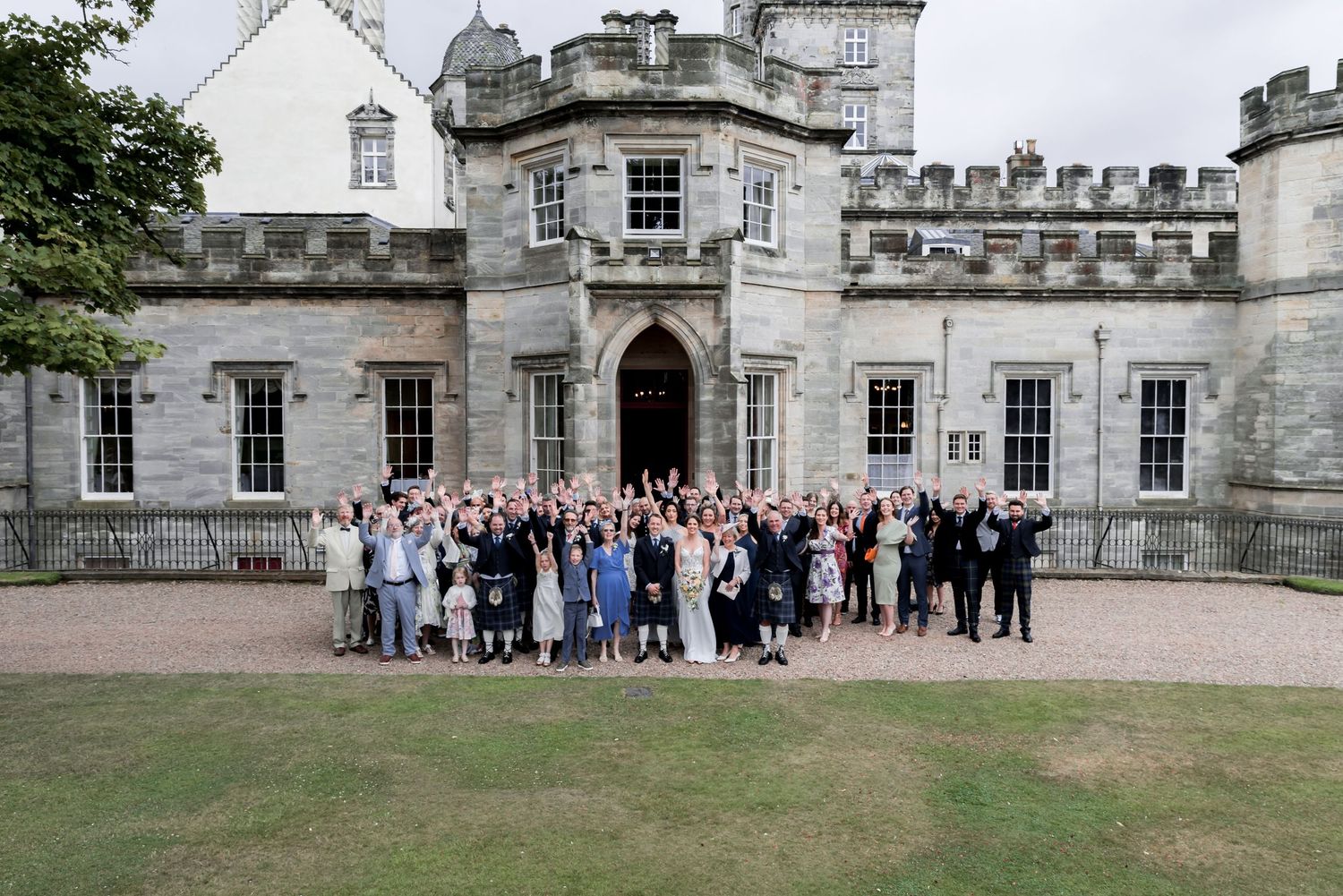 Large group of wedding guests gathered outside a grand stone castle with crenellated walls and ornate architecture.