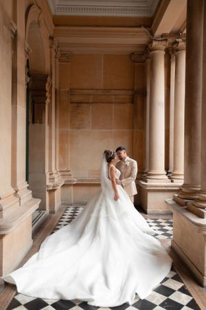 Bride and groom embracing in an elegant colonnade with checkered marble floors and warm stone pillars.