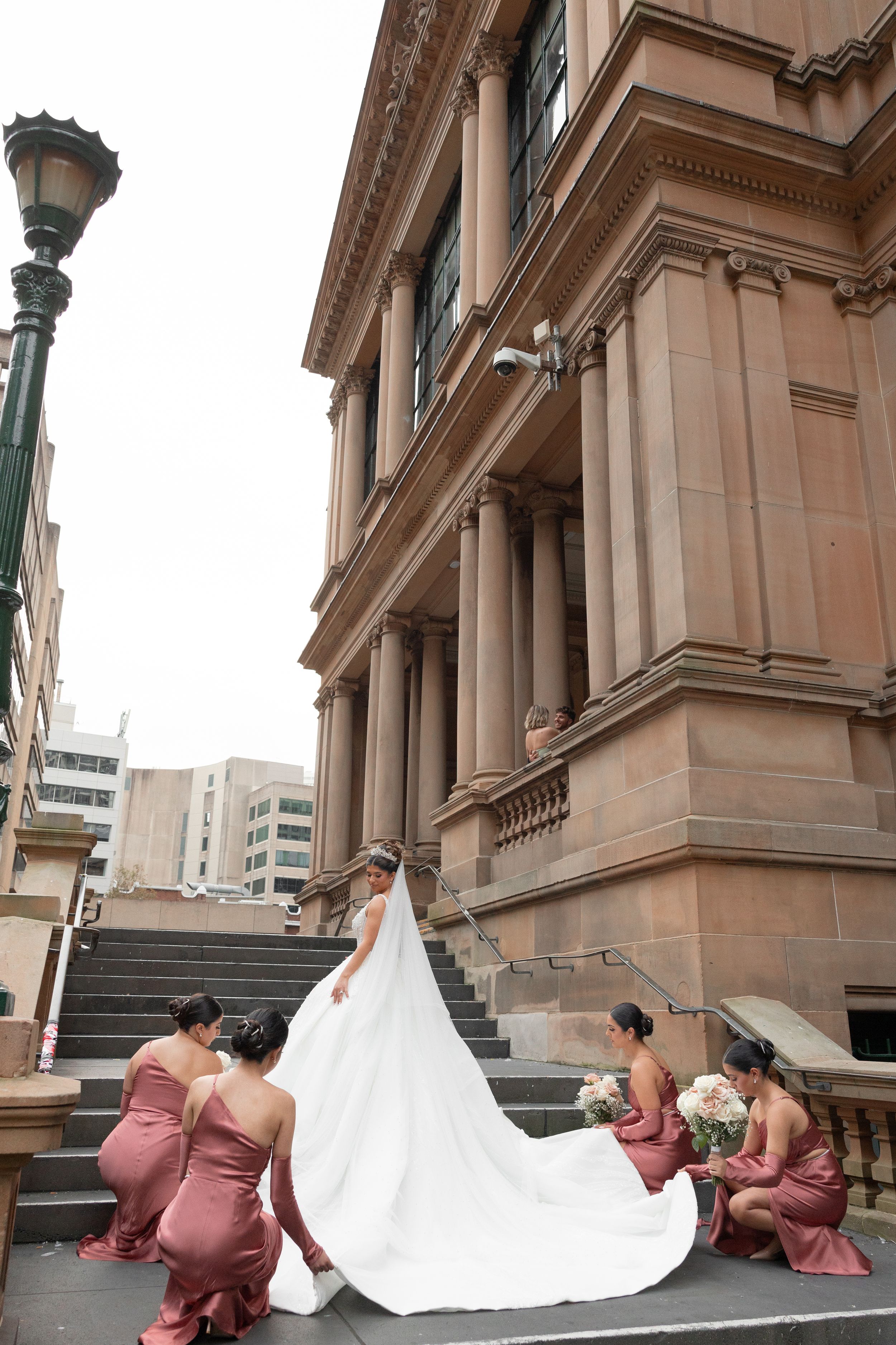 Bridesmaids in pink satin dresses arranging bride's cathedral train on steps of a grand building.