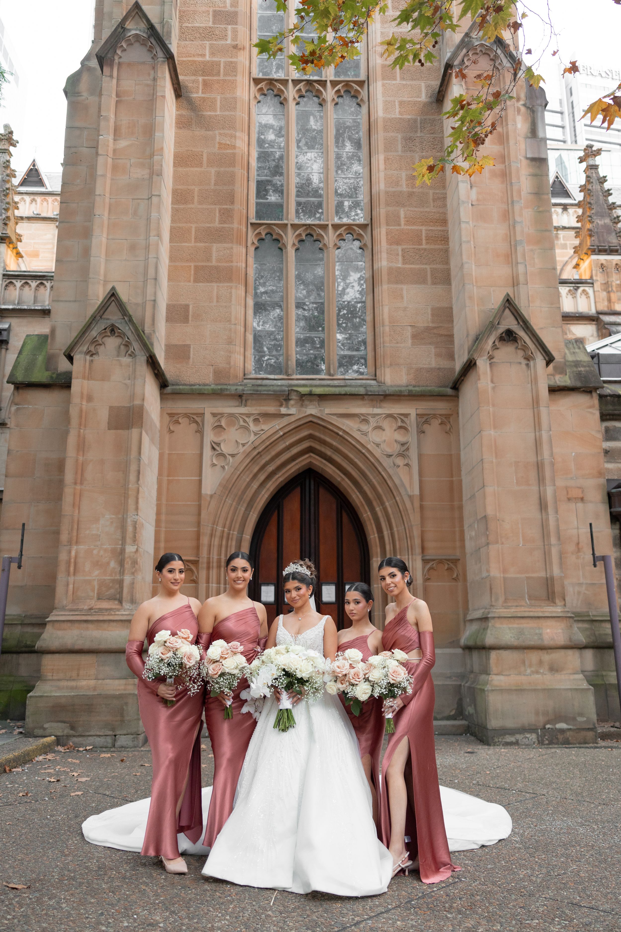 Bride in white gown with four bridesmaids in dusty rose dresses posing before a Gothic stone church.
