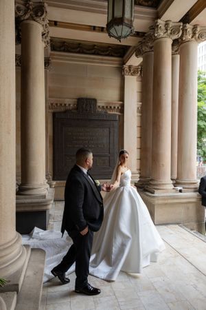 Father walking bride in a voluminous white gown through a classical colonnade with tall stone columns.
