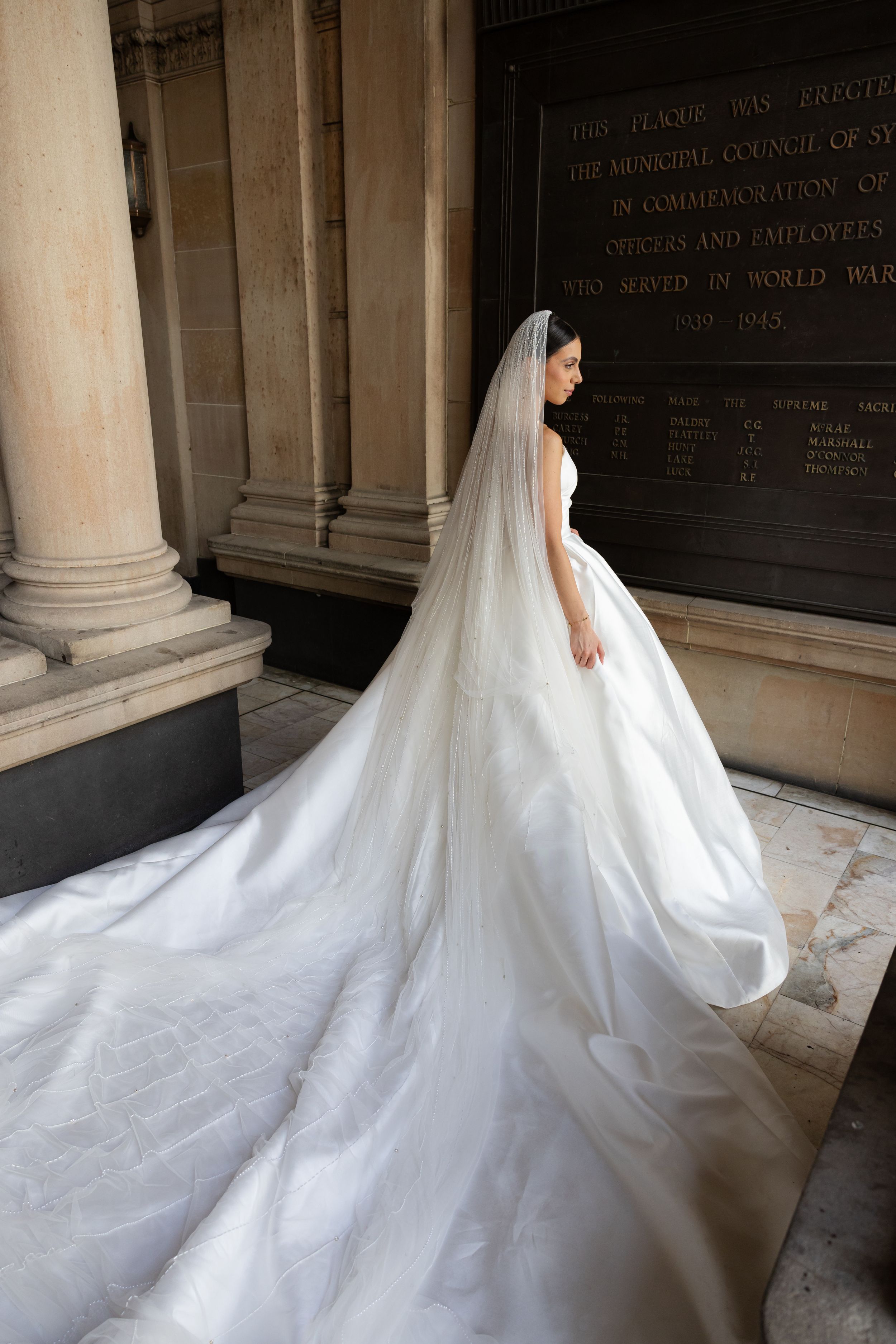 Bride in a stunning cathedral-length veil and ball gown standing beside a stone memorial wall.