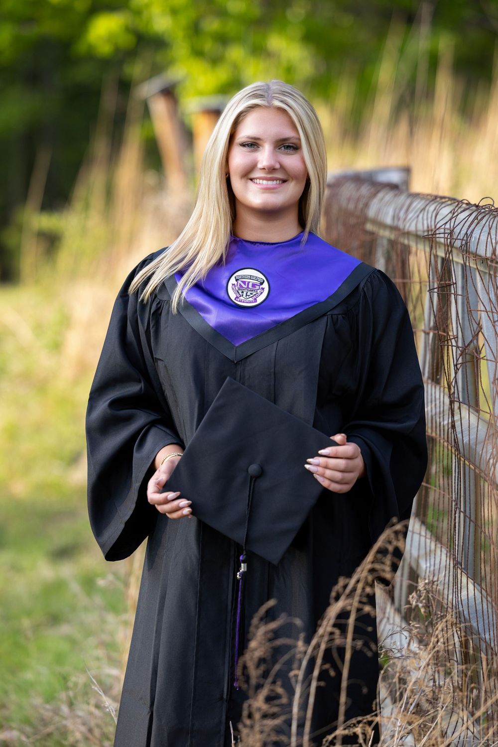 Smiling blonde graduate in black cap and gown with purple hood poses outdoors near a fence.