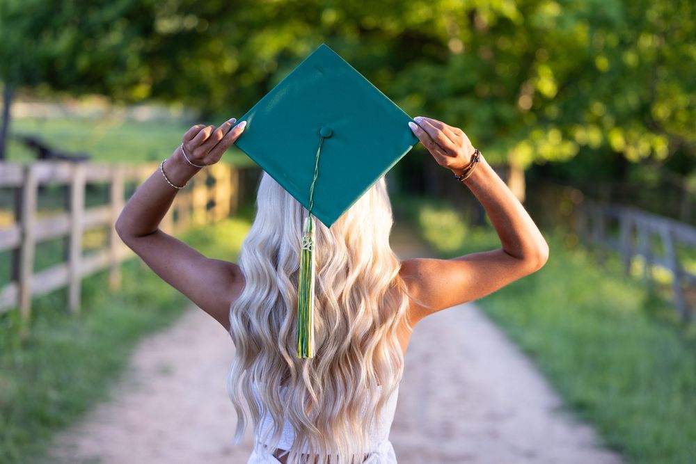 Blonde graduate holding teal graduation cap overhead on tree-lined pathway outdoors.