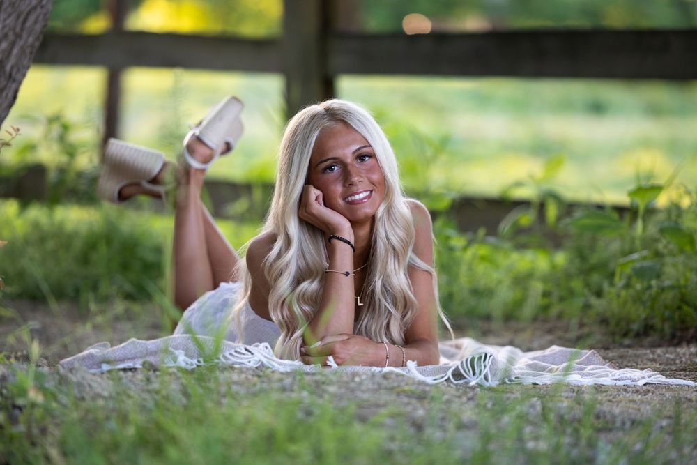 Smiling blonde woman lying on a blanket in a sunny green park, wearing a white dress with heels kicked up.