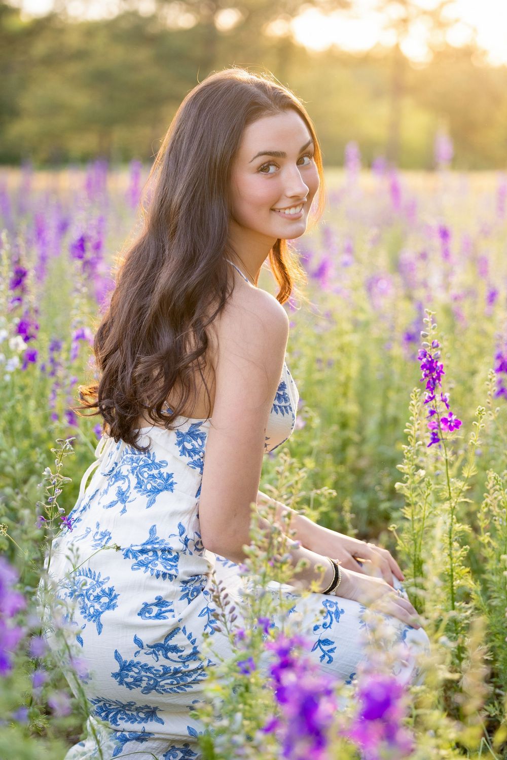 Young woman in blue floral dress smiling in a purple wildflower field at golden hour sunset.