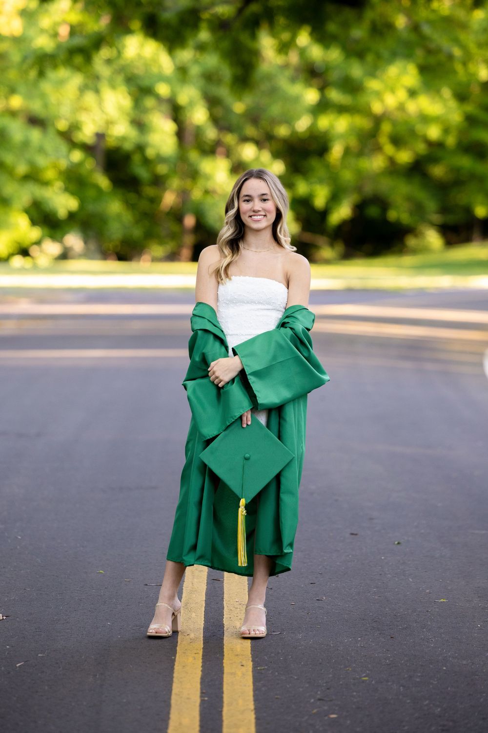 Young woman in white dress holding green graduation cap and gown, smiling on a tree-lined road.