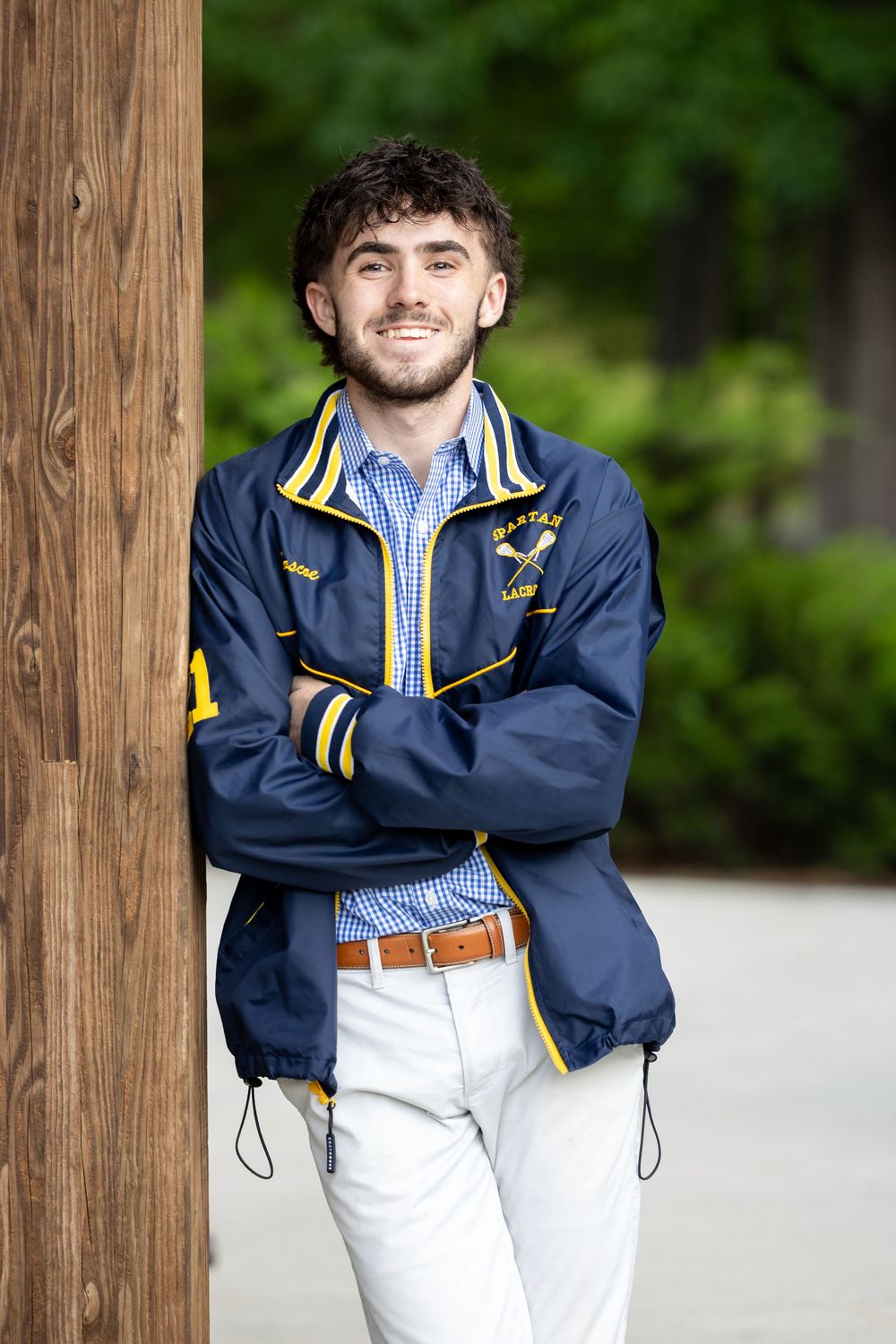 Young man in navy blue and gold varsity jacket with crossed arms leaning against wooden post outdoors.