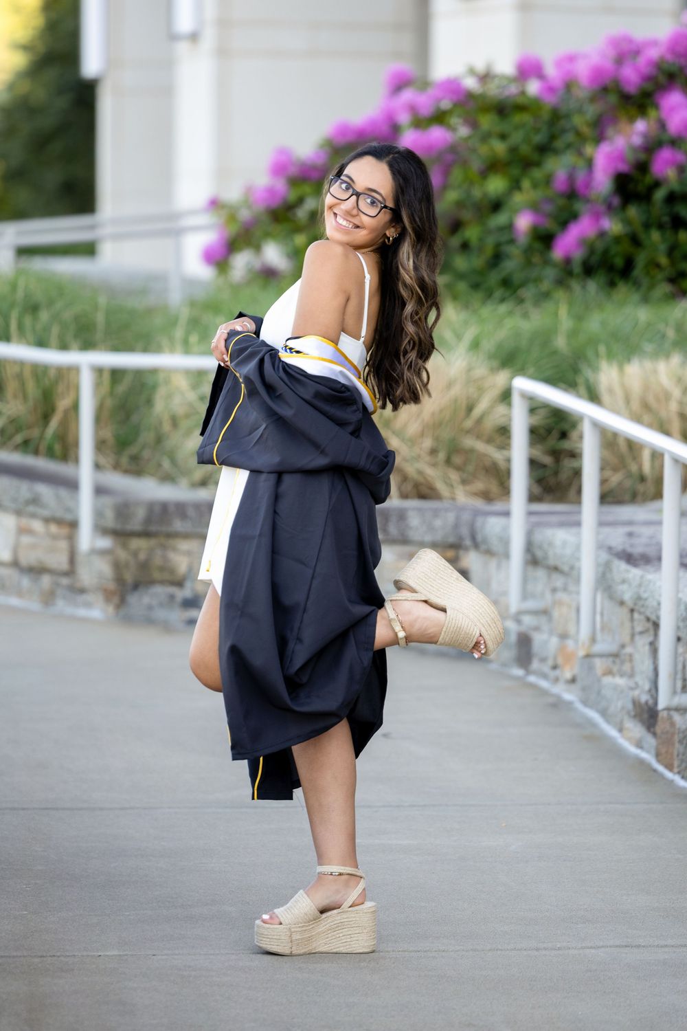 Smiling graduate in black gown holding cap, wearing glasses and wedge sandals, posing outdoors near blooming flowers.