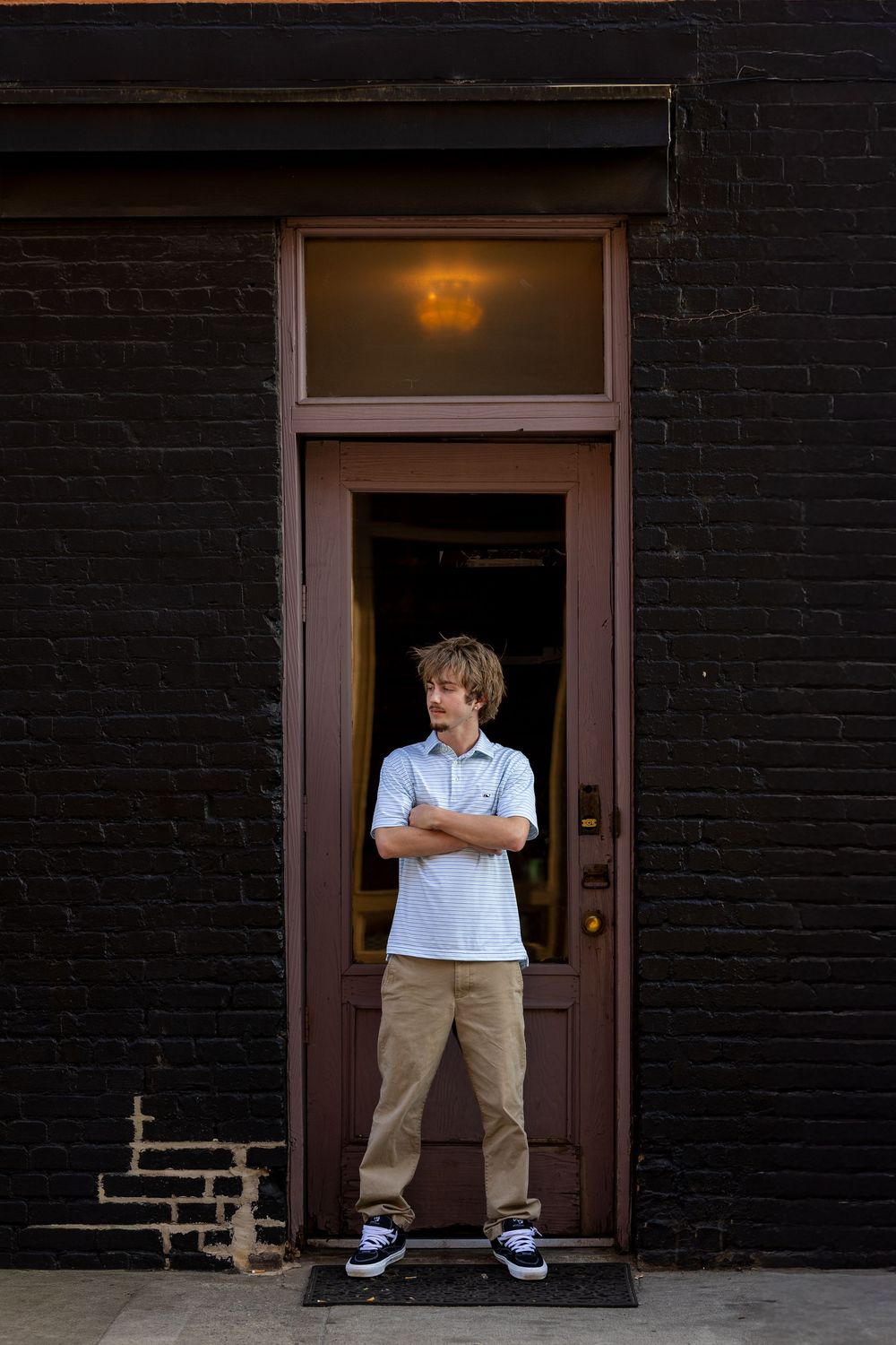 Teen boy in blue shirt and khakis stands arms crossed in front of a dark brick building doorway.