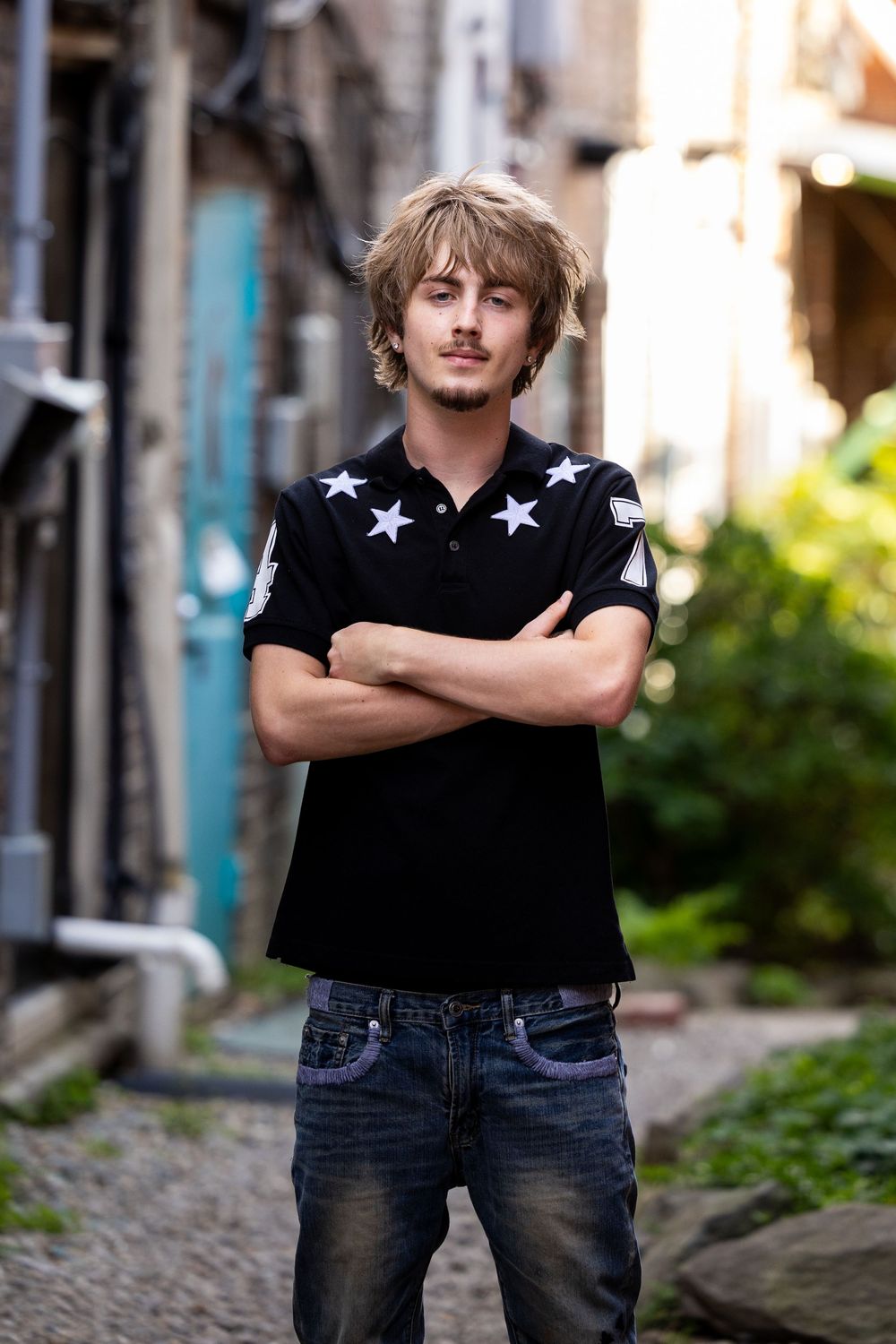 Young man with blonde hair, arms crossed, wearing a black star-print polo shirt and jeans, standing in an urban alley.