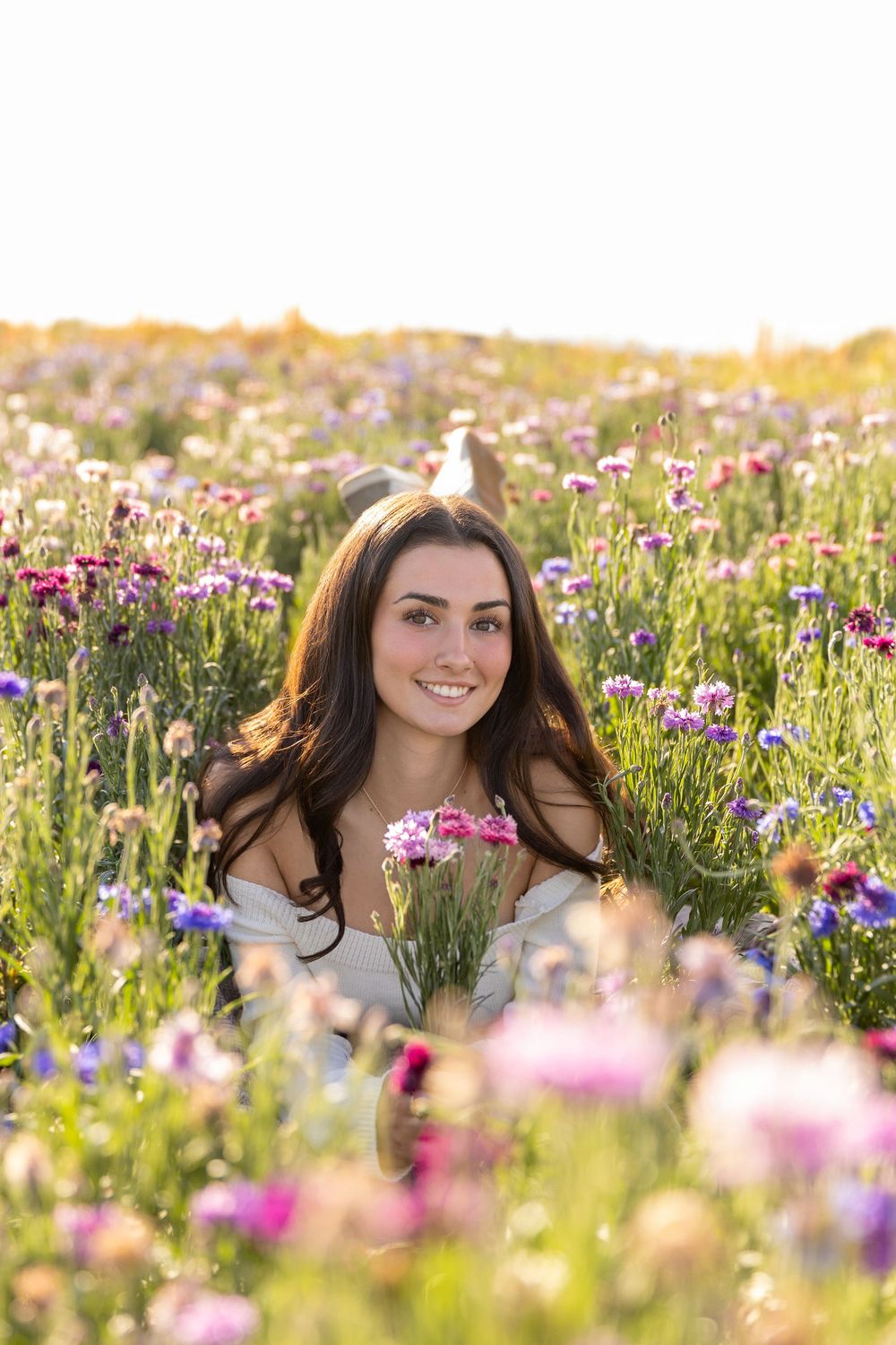 Young woman smiling in a vibrant wildflower field holding pink flowers, bathed in warm golden sunlight.