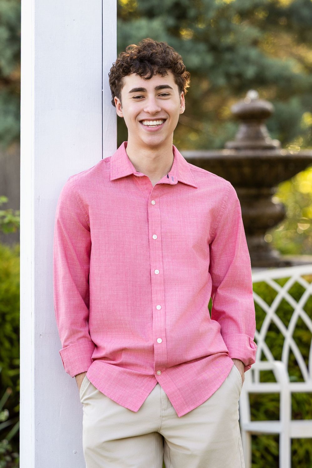 Smiling teenage boy in pink linen shirt and khaki pants leaning against white pillar in garden.