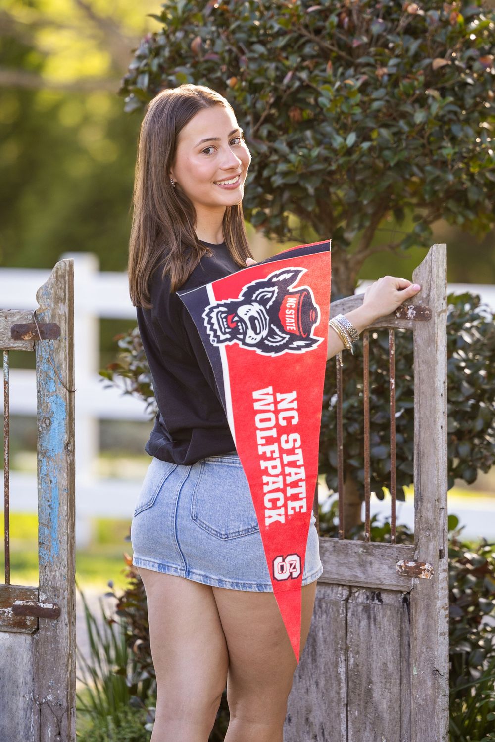 Young woman smiling and holding an NC State Wolfpack pennant flag while leaning against a rustic wooden gate outdoors.