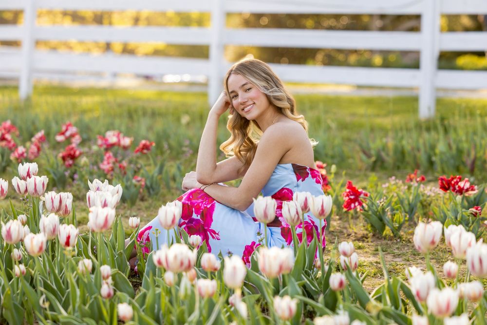 Young blonde woman in blue dress sitting among pink and red tulips by white fence in golden hour light.