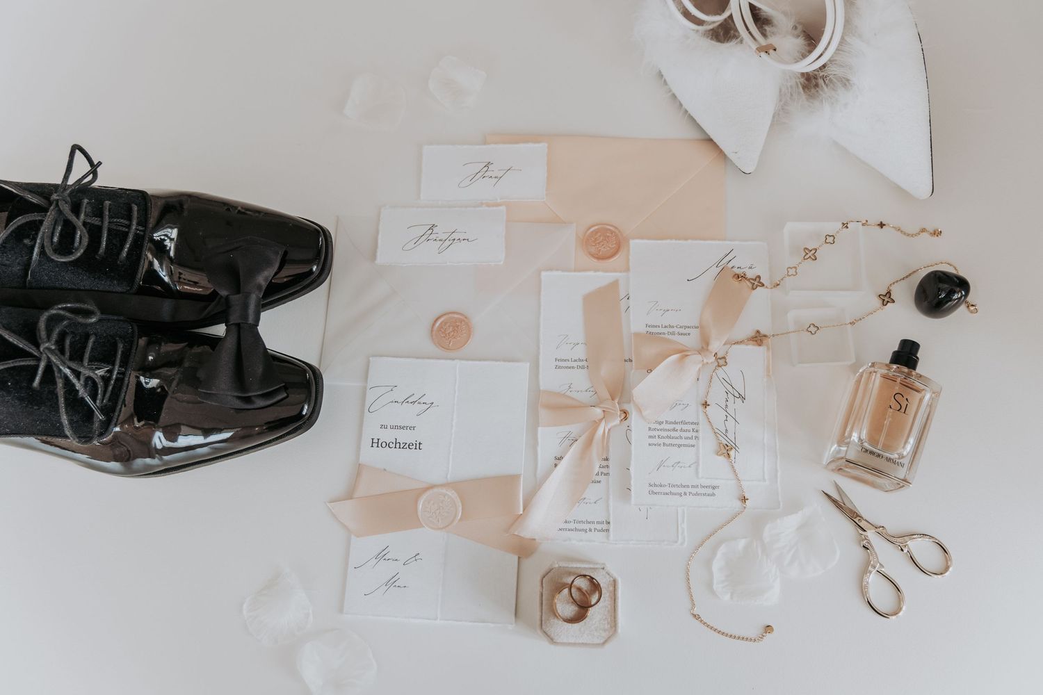 Wedding details flatlay with formal shoes, invitation cards in peach tones, perfume bottle and scissors on white background.
