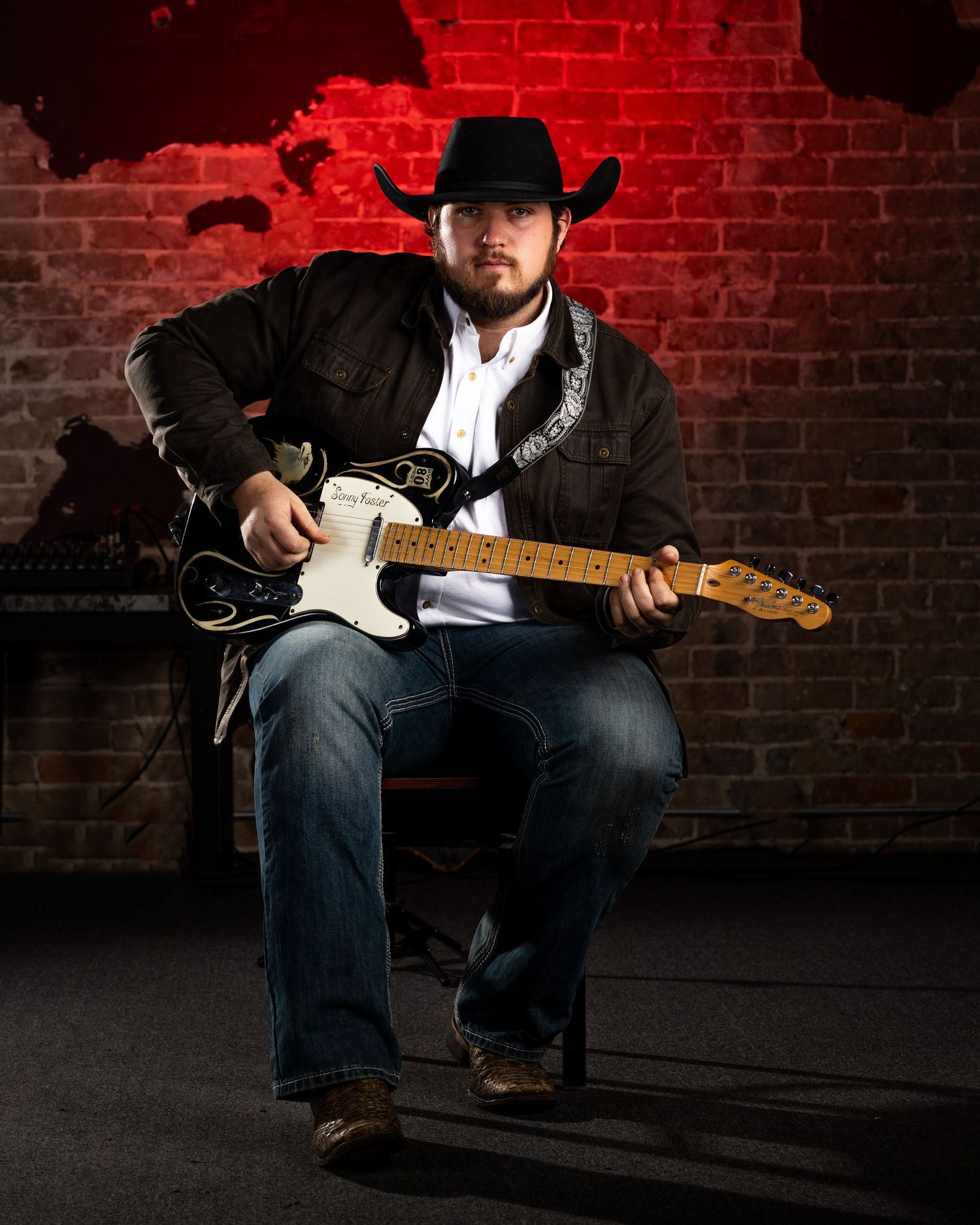 Country musician in black cowboy hat and jacket poses with Fender Telecaster guitar against red brick wall background.