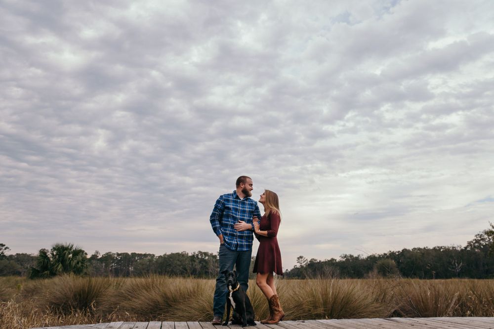 Couple poses for engagement photos against dramatic cloudy sky in an open field.