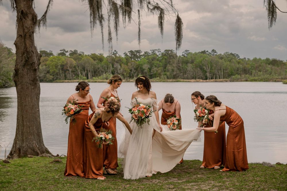 Bridesmaids in rust-colored dresses gather by a lakeside with the bride in white under Spanish moss trees.