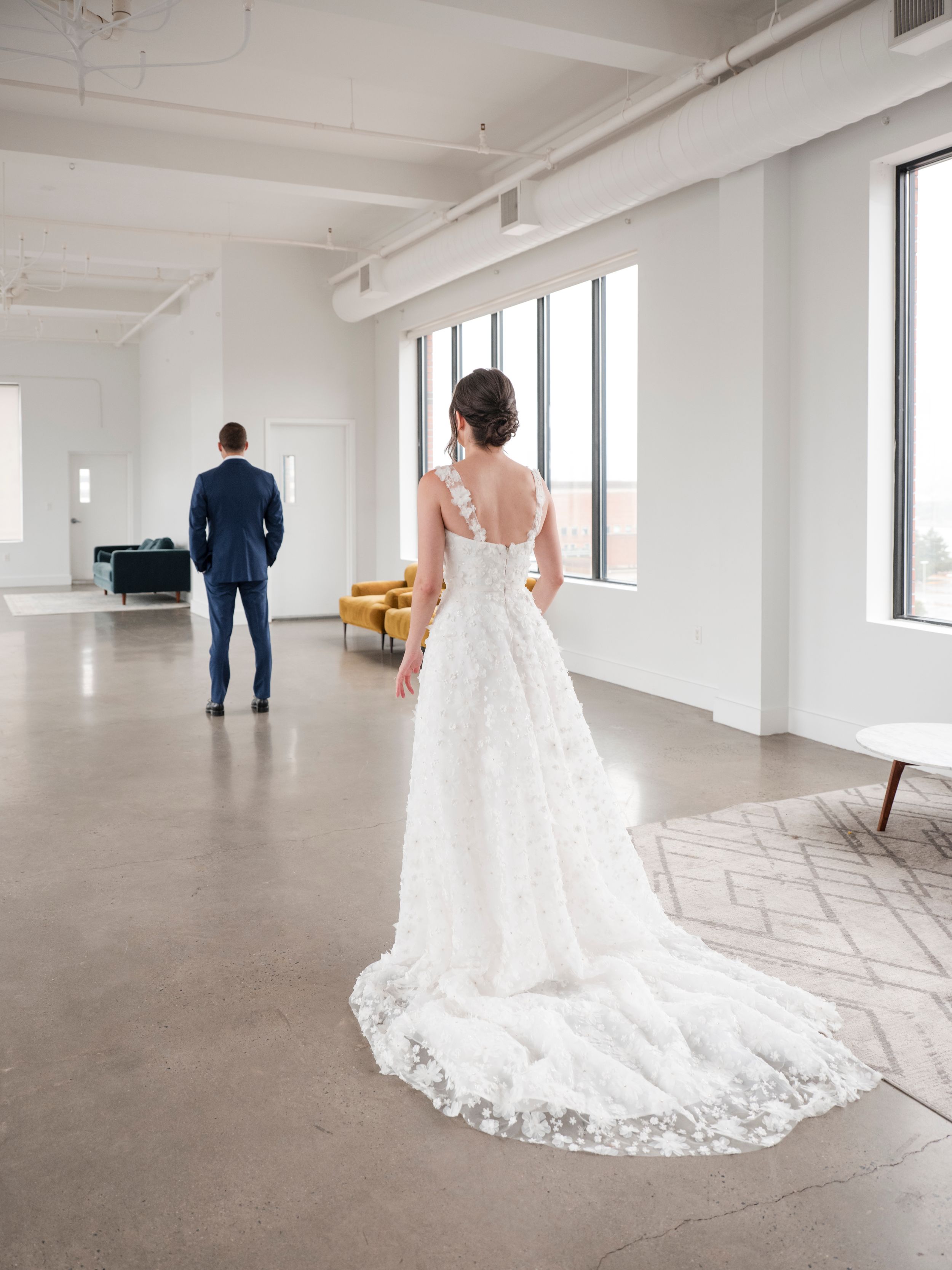 A wedding dress with intricate lace train trails across a modern minimalist room with large windows.