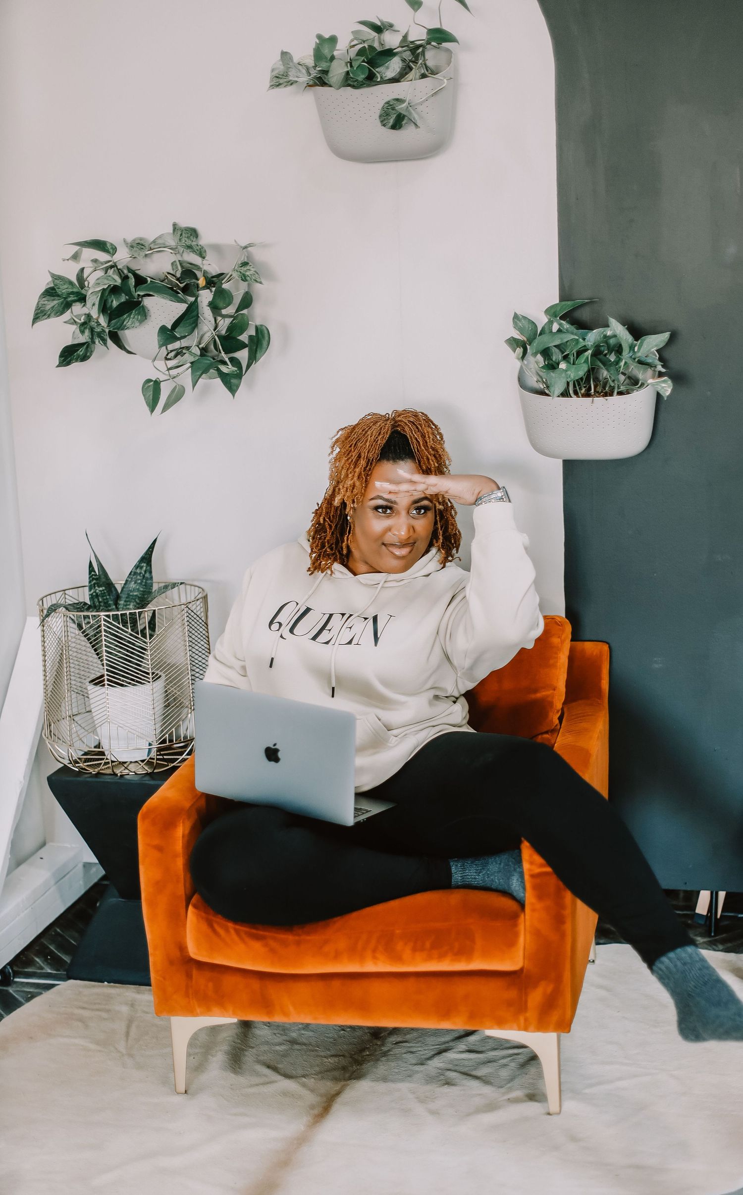 Lifestyle photo working on laptop in orange chair with hanging plants on wall.