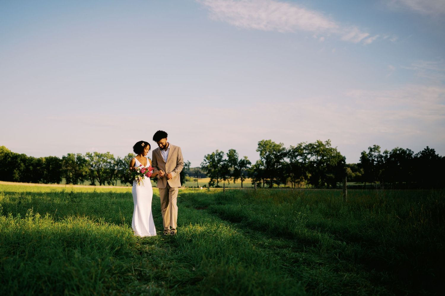 A couple stands together in a sunlit meadow during sunset with trees in the background.