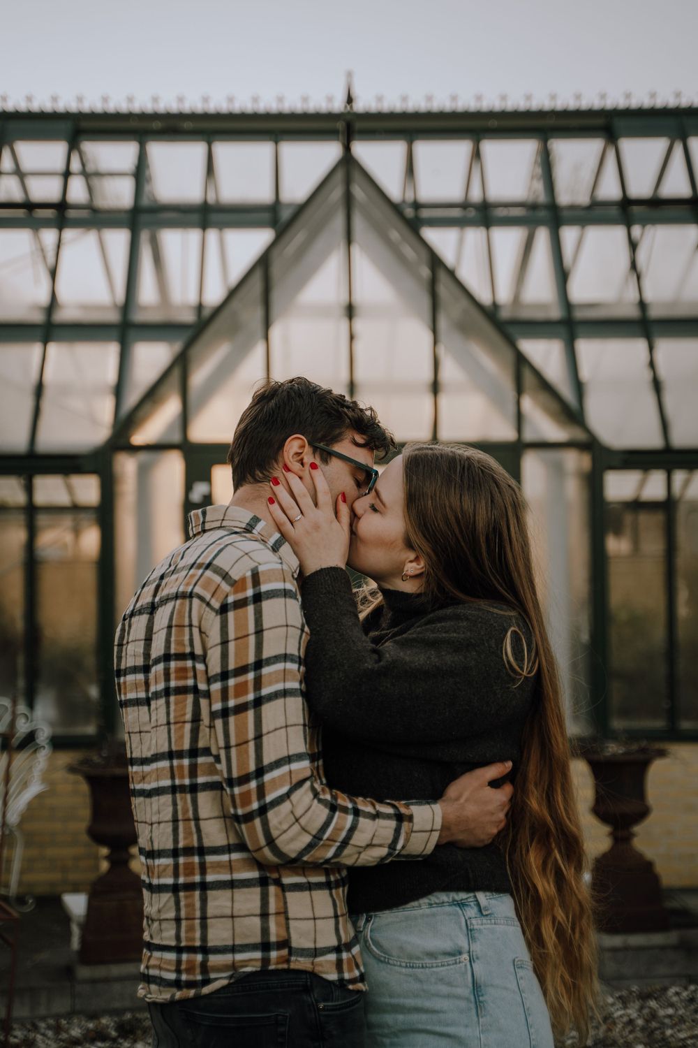 A couple shares a romantic kiss against the backdrop of a greenhouse's geometric architecture during golden hour.