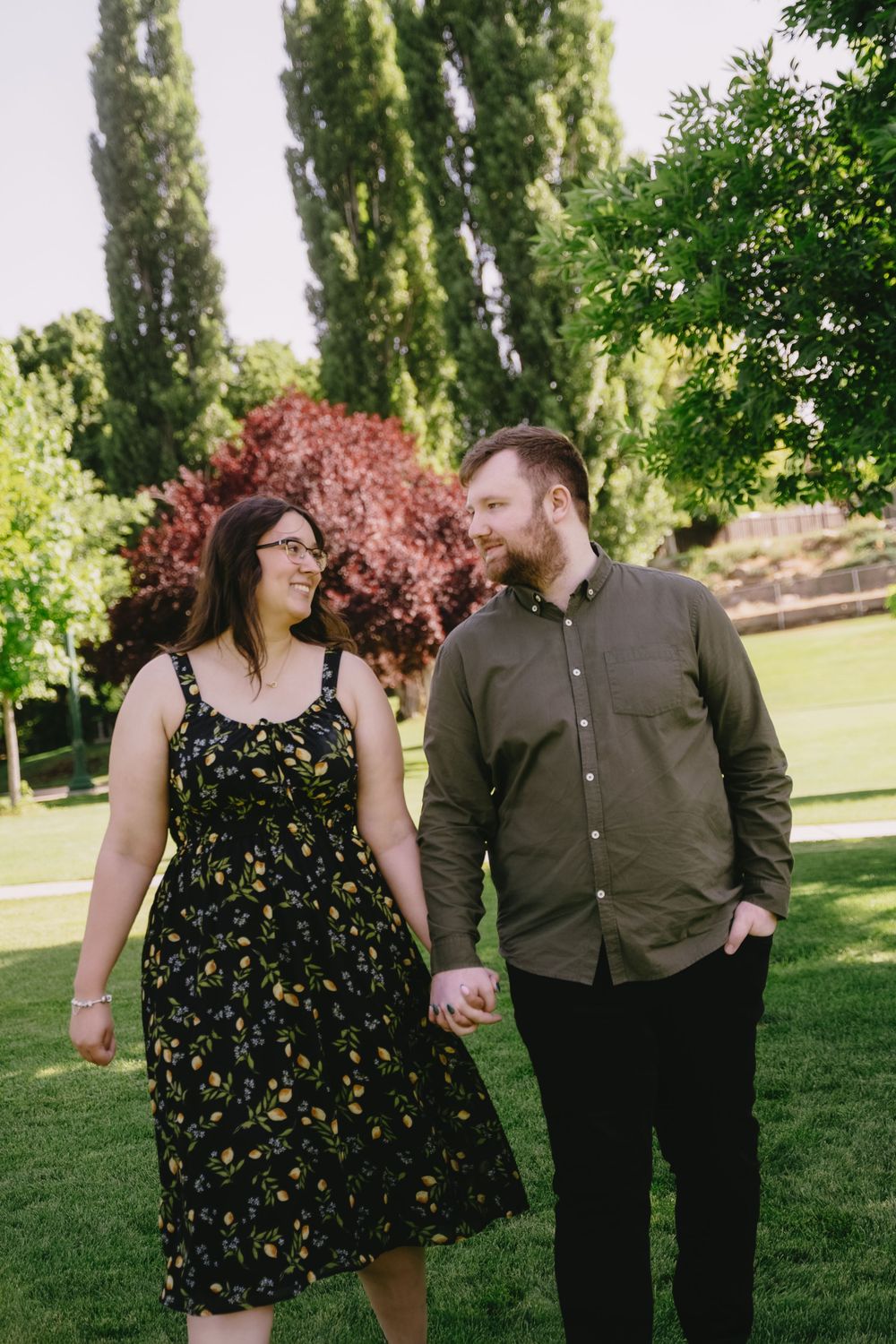 A couple shares a romantic moment together in a lush green park with tall poplar trees and colorful foliage in the background.