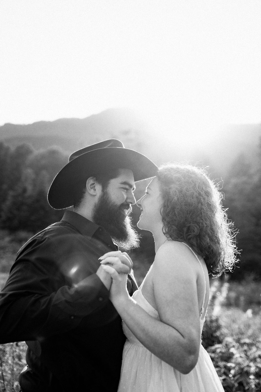A romantic black and white portrait of a couple embracing outdoors in sunlight with mountains in background.