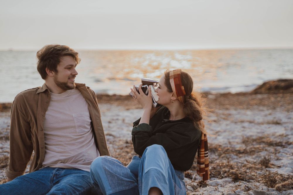 Two people sitting on rocky beach at sunset while one takes pictures with a camera.