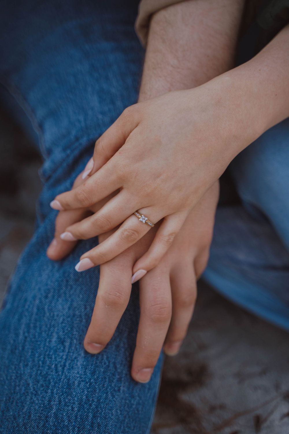 Close-up of hands with a diamond engagement ring resting on blue denim jeans.