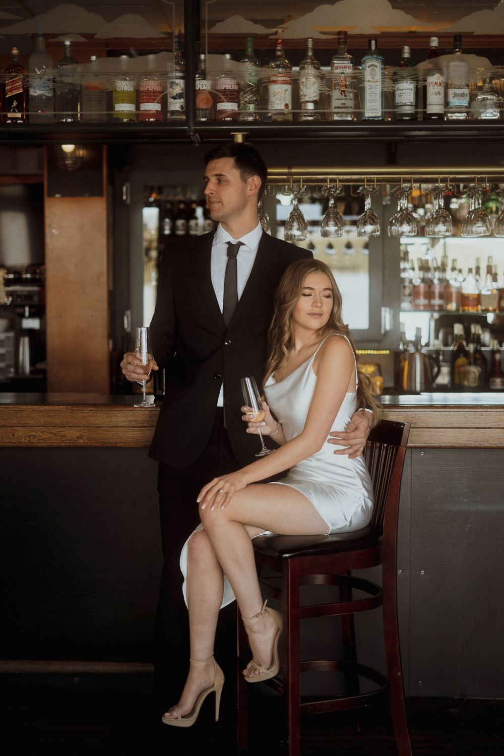 A couple in formal attire poses elegantly at an upscale bar with bottles displayed on shelves behind them.