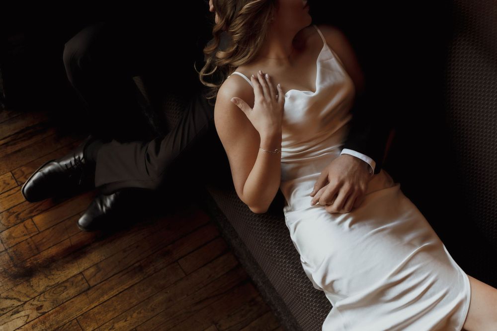 A minimalist wedding dress catches soft light while draped across a dark leather couch on hardwood floors.