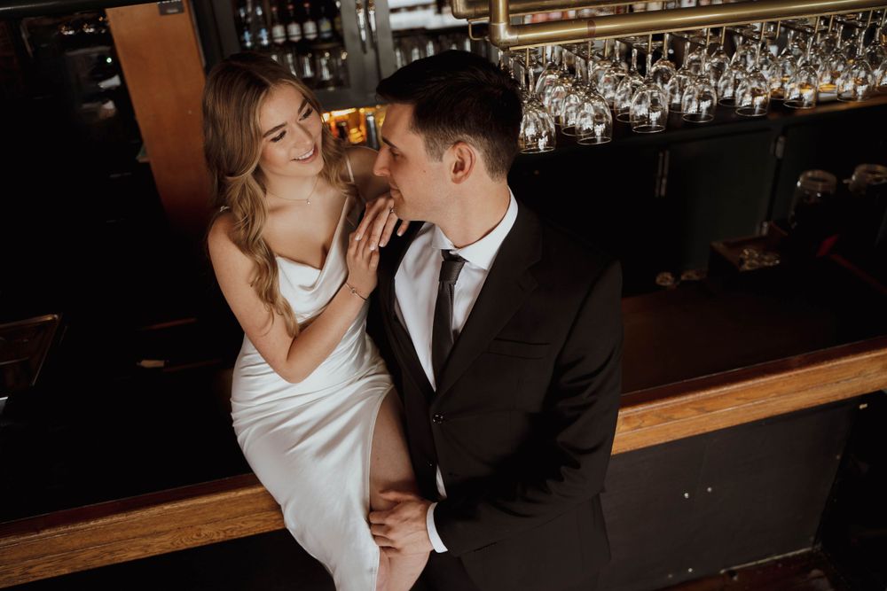 A couple in elegant wedding attire shares an intimate moment at the bar counter with wine glasses hanging overhead.